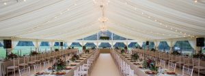 Long wedding reception tables set up inside a large white marquee with chandeliers and string lights.