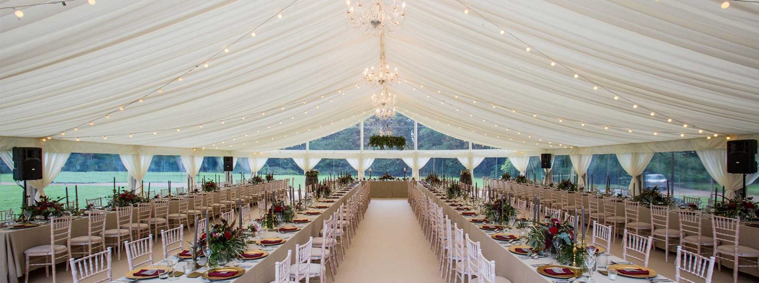 Long wedding reception tables set up inside a large white marquee with chandeliers and string lights.